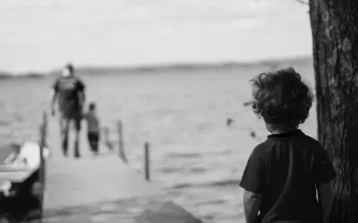 Bambino con capelli ricci guarda verso un molo sfocato dove camminano due figure. Un senso di isolamento, quasi un attaccamento disorganizzato, aleggia nell'aria.