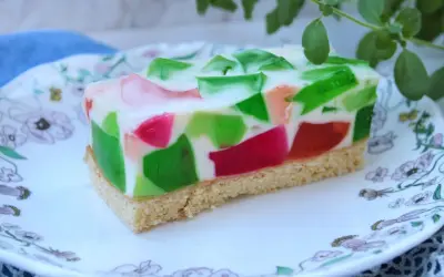 Kolorowe ciasto z galaretkami w kostkę, z zielonymi i różowymi kawałkami zatopionymi w białej masie, na spodzie biszkoptowym.