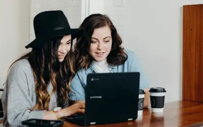 Två unga kvinnor diskuterar sin skuldkvot och bolån framför en laptop, med kaffekoppar på bordet.