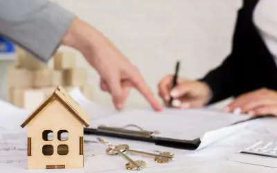 Un perito per valutazione casa indica un documento mentre un'altra persona firma. Chiavi e modellino di casa sul tavolo.
