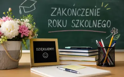 Kwiaty, tabliczka z napisem "DZIĘKUJEMY!" i uśmiechem, stos książek i przybory piśmiennicze na biurku. Na tablicy napis "ZAKOŃCZENIE ROKU SZKOLNEGO".