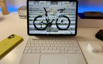 Na ekranie iPada z systemem ipados 26 widoczny jest rower szosowy z napisem "witamy". Obok leży żółty iPhone i biała klawiatura.