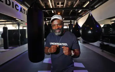 A man in a "UNDEFATED" shirt and Mayweather Boxing + Fitness hat poses with fists up in a gym, ready for Floyd Mayweather training.