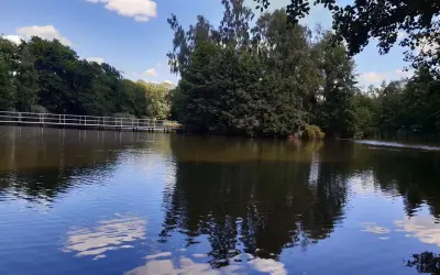 Spokojne jezioro z odbijającymi się chmurami i drzewami. W oddali biały mostek, idealne miejsce na wypoczynek w Golina ośrodek wypoczynkowy.