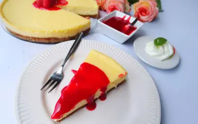 Ein Stück New York Cheesecake mit roter Fruchtsauce und Himbeeren, daneben eine kleine Schüssel Sauce und ein Klecks Sahne.