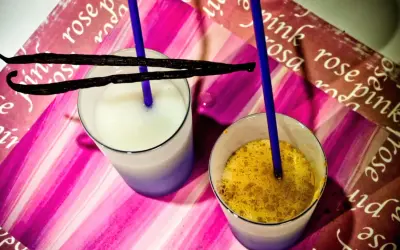 Due bicchieri con latte di riso, uno bianco e uno con spezie dorate, adornati da baccelli di vaniglia e cannucce blu su sfondo rosa.