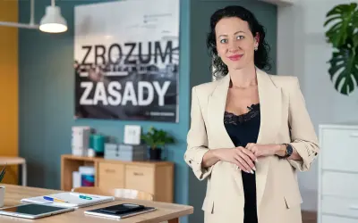 Kobieta w beżowej marynarce, obok niej na biurku leży tablet i notatnik, który może służyć jako księga przychodów i rozchodów.