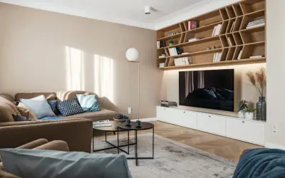 Przytulny salon z sofą i regałem, gdzie dominują kolory pasujące do beżu i brązu. Jasne ściany i drewniane elementy tworzą ciepłą atmosferę.