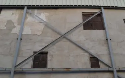 Estructura metálica en forma de X que refuerza una pared de concreto con ventanas de madera. La riostra, en este caso, es un elemento de soporte crucial.