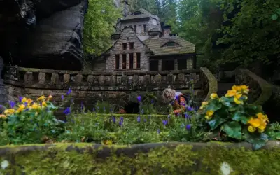 Kamienny domek w skalnym otoczeniu, idealne miejsce na czeska szwajcaria noclegi. Wokół kwitnące kwiaty i zielone drzewa.