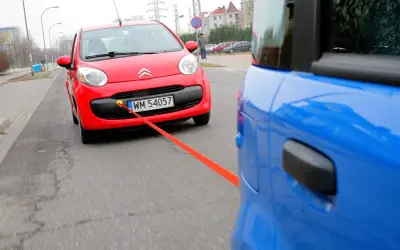 Czerwony Citroen holowany przez niebieski samochód. Pomarańczowa linka holownicza jest napięta, sugerując ruch i prędkość.