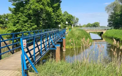 Błękitny mostek dla rowerzystów i pieszych nad kanałem na Żuławach Wiślanych. W tle widać kolejny most i bujną zieleń.