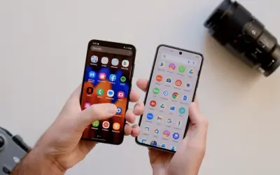 Dwa telefony w dłoniach, jeden z ikoną aparatu, drugi z ikoną aparatu. Który telefon ma lepszy aparat? Obok leży obiektyw aparatu.