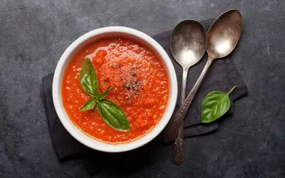 Schüssel mit bester Tomatensauce, garniert mit Basilikum und Pfeffer, daneben zwei Löffel auf dunklem Hintergrund.