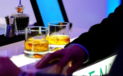 Dwa kieliszki złocistej whisky na barze, idealne na dzień whisky. Dłoń w garniturze sięga po drinka.