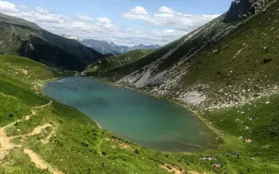 Un sentiero serpeggia tra prati verdi verso il lago Branchino, le cui acque turchesi riflettono le montagne circostanti. Alcune persone si godono un picnic sulle rive.
