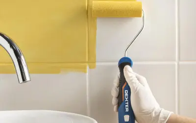Mano con guante blanco pintando azulejos de cocina de blanco a amarillo con rodillo.