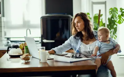 Młoda mama z dzieckiem na rękach pracuje przy laptopie. Czy na macierzyńskim można pracować? Tak, wiele mam łączy opiekę z pracą zdalną.