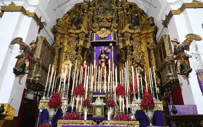 Un altar barroco con imágenes religiosas, velas y flores rojas. Destaca una figura central de Cristo, que parece ser un quinario, rodeado de figuras de la Virgen y otros santos.