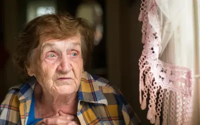 Starsza kobieta zamyślona, z rękami na piersi, przy oknie z koronkową firanką. W jej oczach widać smutek, być może to depresja starcza.