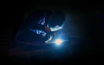 Spawacz w masce ochronnej używa elektrody do żeliwa, tworząc jasny łuk świetlny i dym.