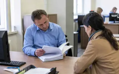Funkcjonariusz w niebieskiej koszuli przegląda dokumenty, siedząc przy biurku naprzeciwko kobiety.