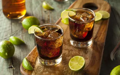 Dwa drinki z colą i lodem, ozdobione ćwiartkami limonki. W tle butelka z bursztynowym płynem. To klasyczny rum z colą, czyli co to jest ron.