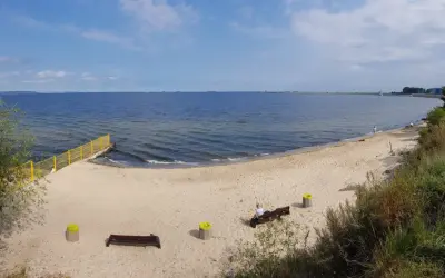 Plaża dla psów z łagodnym zejściem do morza. Na piasku ławki i kosze. W oddali widać falochron.