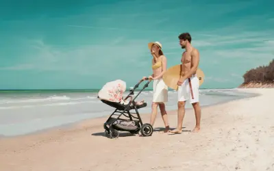 Para spaceruje po plaży z wózkiem Kinderkraft Askoy. Mężczyzna niesie deskę surfingową.