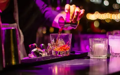 Barman nalewa bursztynowy cordial drink do szklanki z lodem. W tle kolorowe światła.