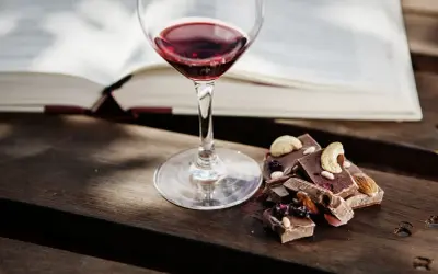 Un bicchiere di vino rosso e cioccolato con frutta secca e noci, un perfetto abbinamento vino e cioccolato, accanto a un libro aperto.