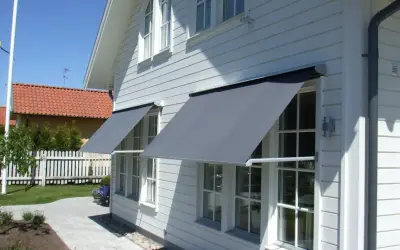 Grå markiser monterade på ett vitt trähus. Solen skiner och skapar skuggor på fasaden.