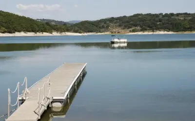 Un pontón blanco navega en el tranquilo embalse de Prada, con un muelle de madera en primer plano y colinas verdes al fondo.