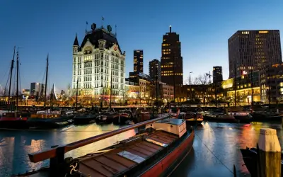Wieczorem w porcie, gdzie jest Rotterdam. Widać zabytkowy budynek, nowoczesne wieżowce i zacumowane łodzie.
