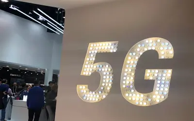 Wielkie, podświetlane litery "5G" na ścianie, symbolizujące przyszłość technologii. W tle widać ludzi i stoiska targowe.