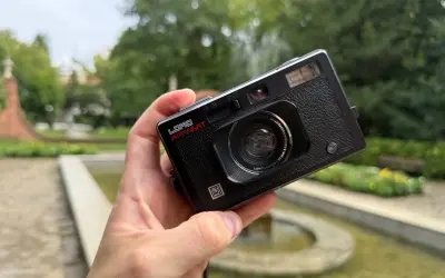 Czarna, analogowa lomografia LomoApparat z obiektywem 21mm. W dłoni, na tle zieleni i fontanny.