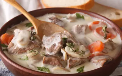 Un plat réconfortant de véritable recette blanquette de veau, avec des morceaux de viande tendre, des champignons et des carottes dans une sauce crémeuse, servi dans un bol en bois avec une cuillère.
