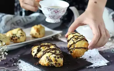 En hand plockar upp en chokladscone, dekorerad med chokladglasyr. Fler chokladscones ligger på ett fat.