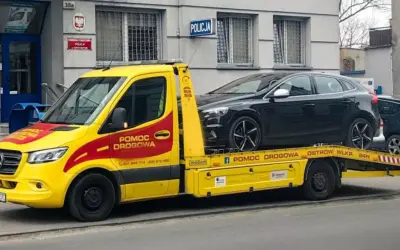 Żółta laweta z napisem "Pomoc drogowa" przewozi czarny samochód przed budynkiem policji. Konfiskata samochodu w akcji.