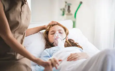 Dziewczynka w szpitalnym łóżku z maską tlenową, walcząca z hipoksją. Opiekunka czule kładzie dłoń na jej głowie.