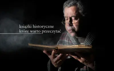Starszy mężczyzna w okularach pochyla się nad starą księgą, zanurzając się w lekturze. Wokół unosi się pył, podkreślając atmosferę tajemnicy i wiedzy zawartej w książkach historycznych.