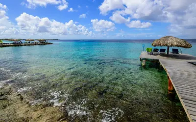 Bonaire wyspa: drewniany pomost z leżakami i parasolem, turkusowa woda, skaliste dno, chmury na niebie.
