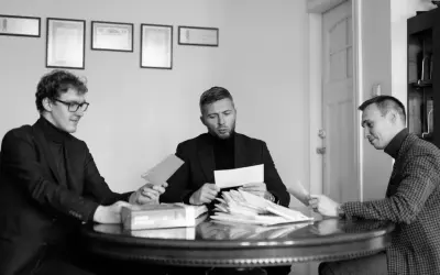 Trzech mężczyzn przy okrągłym stole analizuje dokumenty. Jeden z nich trzyma kopertę, jakby właśnie otrzymał wyrok w zawieszeniu.