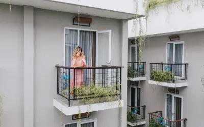 Kobieta w różowej sukience stoi na balkonie, opierając się o balustradę. Wokół rosną zielone rośliny, tworząc naturalną zasłonę na balkon.
