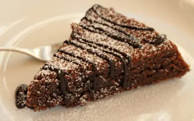 Una fetta di torta caprese al cioccolato, cosparsa di zucchero a velo e con una colatura di cioccolato fondente.
