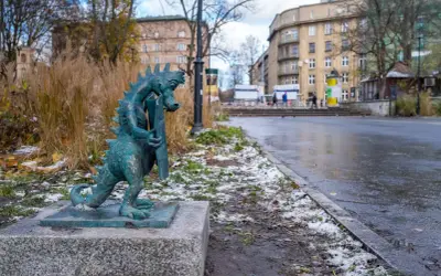 Mały smok z brązu, symbol smoczy szlak kraków, stoi na kamiennym postumencie. W tle miejski krajobraz z budynkami i drzewami.
