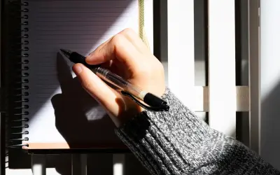 Dłoń pisze w notatniku, planując letnie podróże. Cień pada na kartki, jakby to były święta w lipcu.