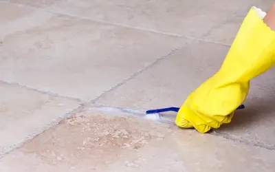 Mano con guanto giallo pulisce le fughe dei pavimenti con uno spazzolino e schiuma.