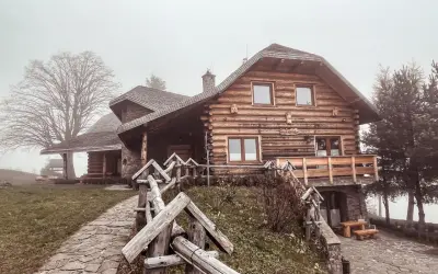 Drewniany dom z kamienną podstawą, kamienista ścieżka i płot. Mglisty dzień dodaje uroku temu miejscu, które oferuje liczne laskowa atrakcje.