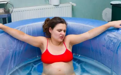 Kobieta w czerwonym bikini przechodzi poród w wodzie. Woda delikatnie faluje wokół niej, a jej twarz wyraża wysiłek.